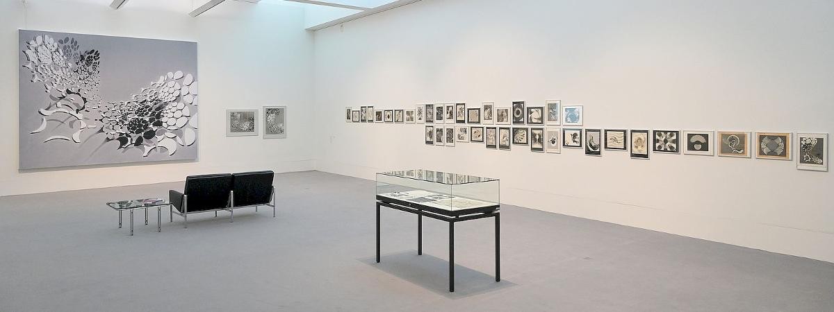 Ausstellungsansicht "Reiner Michelfelder – ars dynografia. Experimentelle Fotografie der 1960er und 1970er Jahre", Foto: Stadt Kornwestheim. Ausstellungsansicht der Ausstellung „Reiner Michelfelder – ars dynografia“ mit einer großformatigen Schwarzweißarbeit links sowie einer langen Reihe abstrakter Fotografien an der Wand. Im Raum befinden sich eine Vitrine und eine Sitzbank .