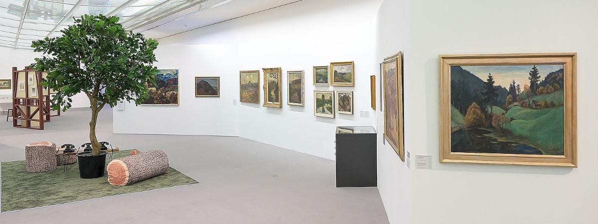Ausstellungsansicht "Stadt – Land – Sehnsucht", Foto: Stadt Kornwestheim. Ausstellungsansicht von „Stadt – Land – Sehnsucht“ mit historischen Landschaftsgemälden an weißen Wänden und einer zentralen Installation aus Baum, Teppich und Sitzmöbeln in Baumstammform.