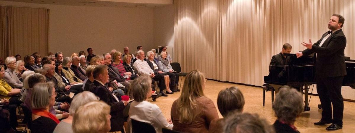 Vernissage mit dem Tenor Reto Raphael Rosin und dem Pianisten Bernhard Epstein im März 2019. Foto: Ingo Kröner. Galerieversammlungssaal mit Sänger, Pianist und Publikum.