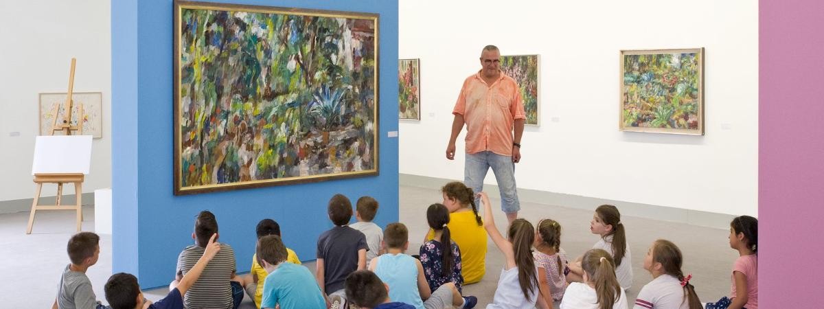 Kinderführung mit Michael Bischof durch die Ausstellung "Manfred Henninger". Michael Bischof steht neben einem Gemälde, auf dem Boden davor sitzen Kinder.