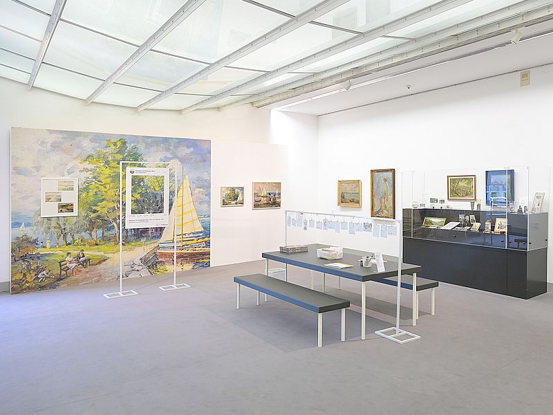 Ausstellungsansicht "Stadt – Land – Sehnsucht", Foto: Stadt Kornwestheim. Ausstellungsansicht der Ausstellung „Stadt – Land – Sehnsucht“ mit Gemälden, Grafiken und Alltagsobjekten. Im Vordergrund stehen Vitrinen und Sitzbänke, im Hintergrund sind großformatige Landschaftsdarstellungen und gerahmte Werke an den Wänden zu sehen.