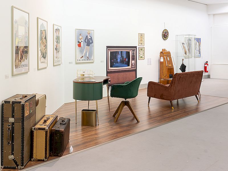 Ein Ausstellungsraum mit Möbeln im Stil der 1950er/1960er Jahre, darunter ein grüner Schreibtisch, ein Sessel, eine braune Couch und Koffer. An den Wänden hängen Werbegrafiken aus dieser Zeit, während ein Fernseher auf einem Holzfußboden platziert ist.