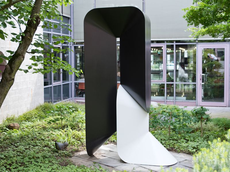 Horst Kuhnert, Raumkörper 74 S.W., 1974/1994. Eine Skulptur von Horst Kuhnert. Eine schwarz-weiße Arbeit aus Polyester. Darum Vegetation. Im Hintergrund der Einblick in das Rathaus.