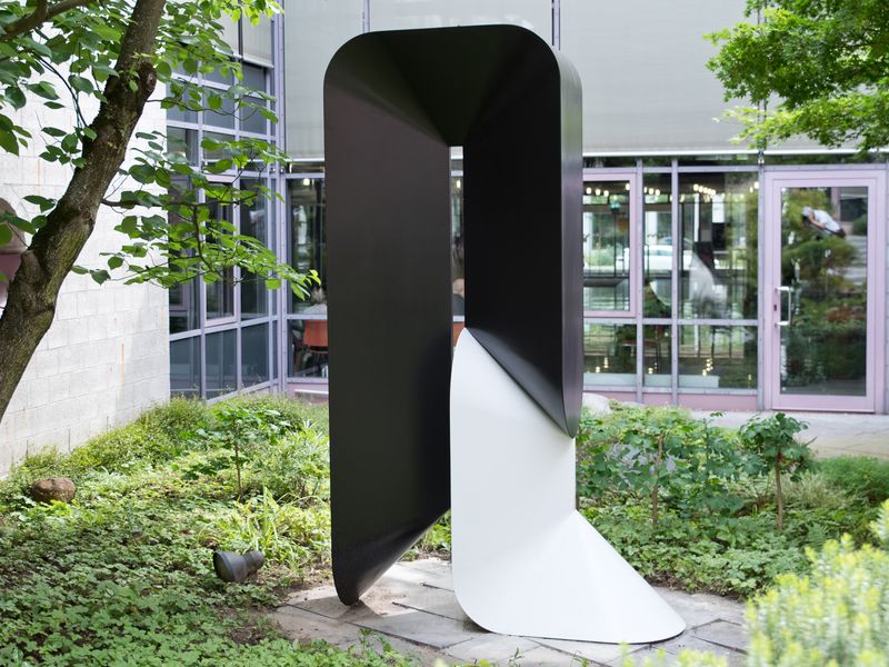 Eine Skulptur von Horst Kuhnert. Eine schwarz-weiße Arbeit aus Polyester. Darum Vegetation. Im Hintergrund  der Einblick in das Rathaus.